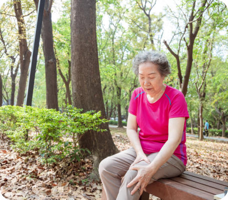 Photo of a senior woman sitting on a bench in a park. She has a pained expression on her face and she is holding her knee.