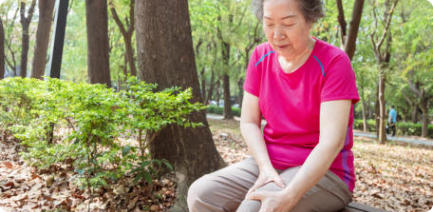 Photo of a senior woman sitting on a bench in a park. She has a pained expression on her face and she is holding her knee.